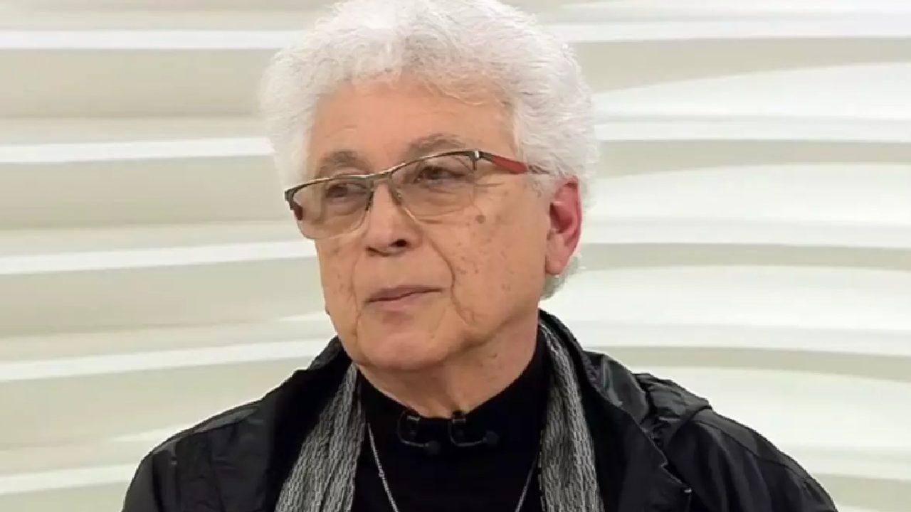 A imagem mostra uma pessoa idosa de cabelos brancos curtos e óculos, usando uma jaqueta preta, cachecol cinza e uma blusa escura. O fundo é claro, com um padrão ondulado branco, típico de um estúdio de televisão