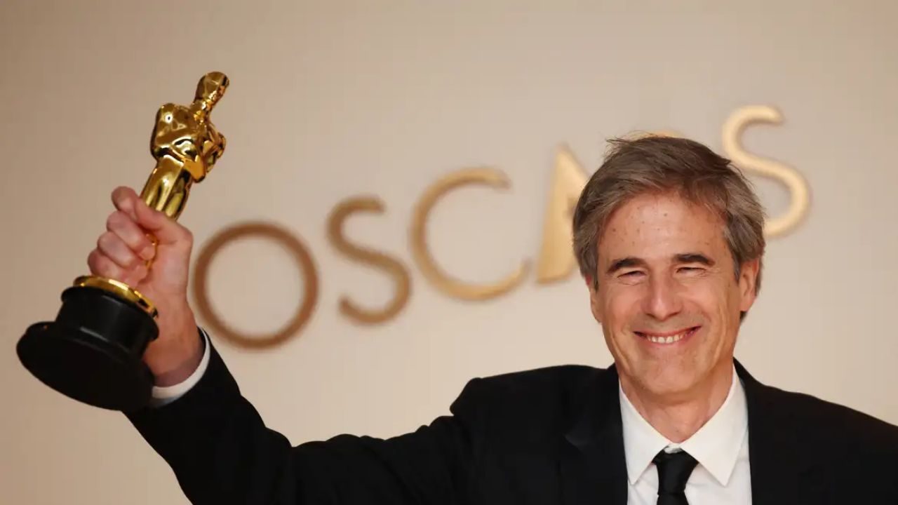 Homem de terno levanta estatueta do Oscar sorrindo diante de painel com nome da premiação