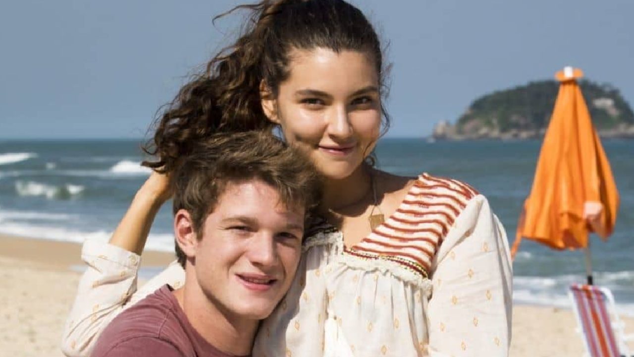 A imagem mostra um casal jovem sorrindo em uma praia. A mulher, com cabelo castanho preso em um rabo de cavalo e usando uma blusa clara com detalhes listrados, está abraçando o rapaz, que tem cabelo curto e veste uma camiseta vermelha