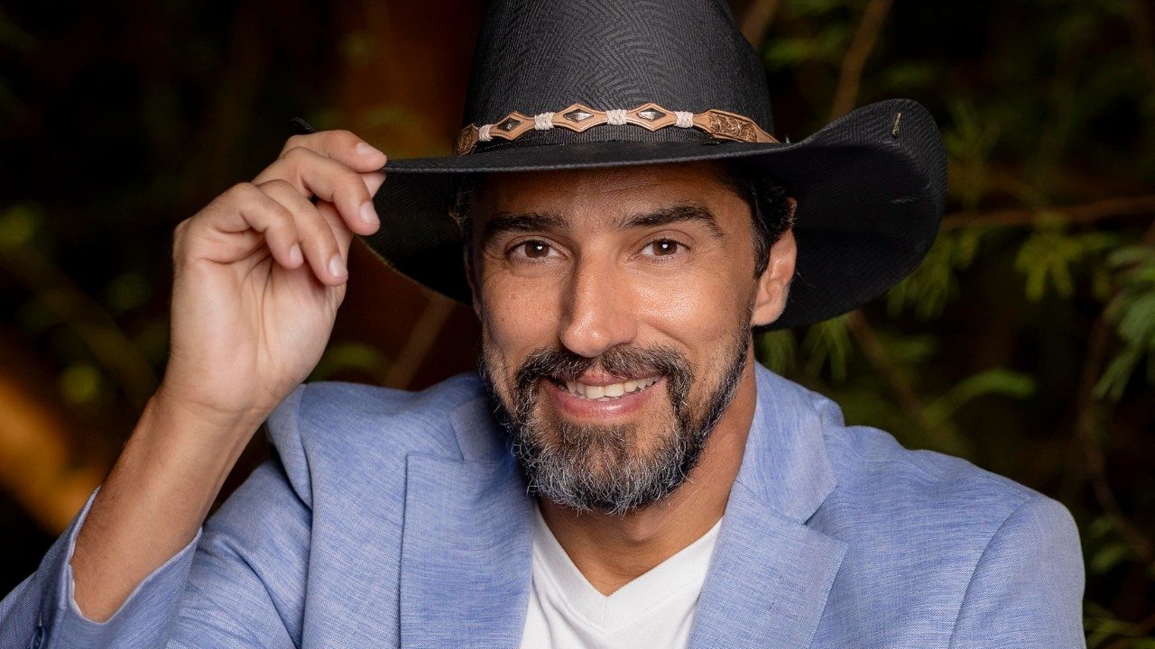 Alberto Cowboy sorri usando chapéu preto e blazer claro enquanto segura a aba do chapéu em ambiente com plantas ao fundo