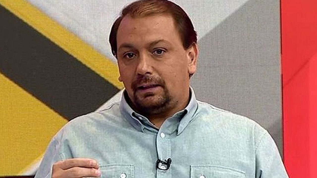 Homem com microfone de lapela fala em estúdio de TV diante de painel com gráfico ao fundo