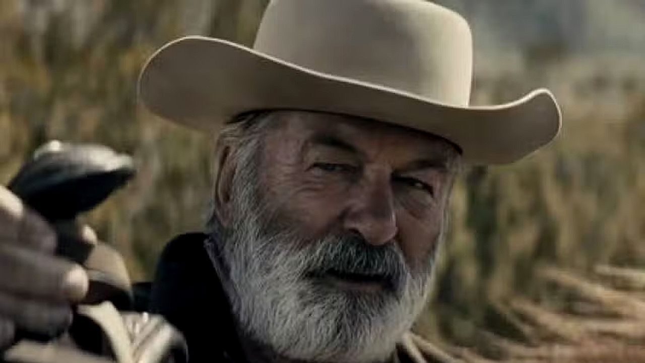 Homem com chapéu de cowboy e barba grisalha aponta arma em cena ambientada no campo