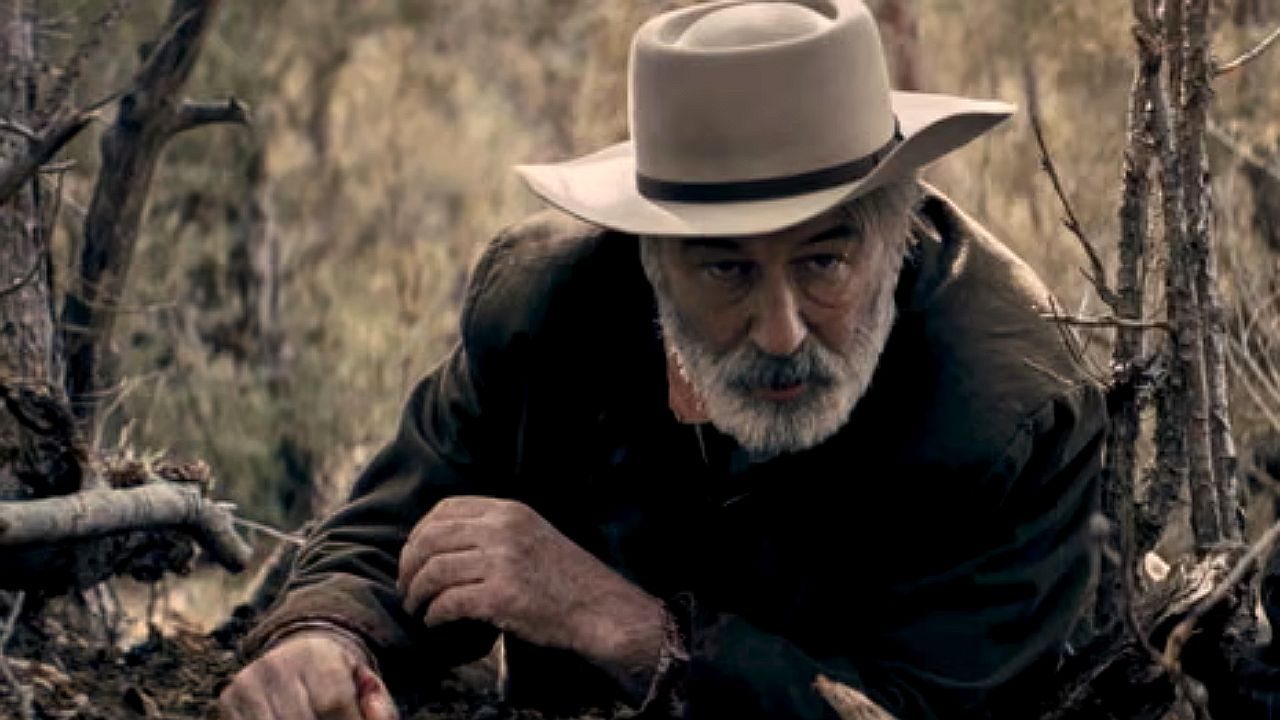 Homem de chapéu e barba observa o chão em área de mata em cena de faroeste