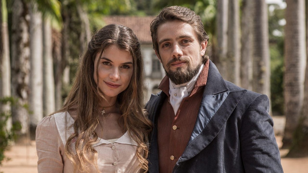 Casal em trajes de época posa junto ao ar livre em cena da novela Além do Tempo.