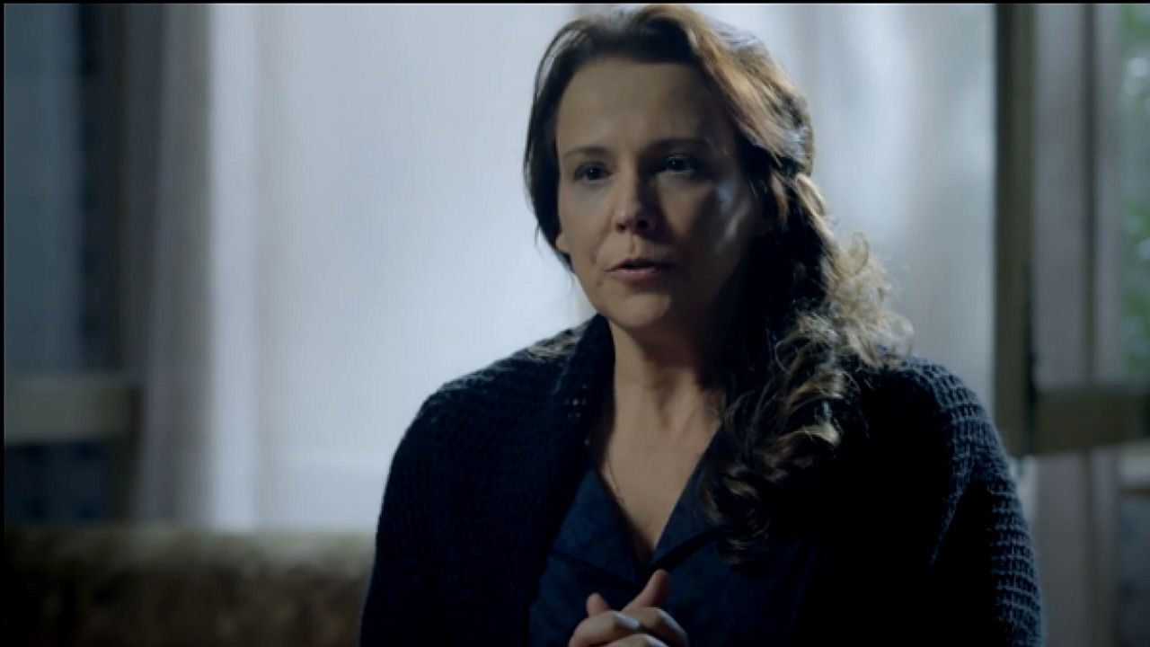 Mulher com expressão séria fala com mãos juntas em ambiente interno com iluminação baixa em cena de novela