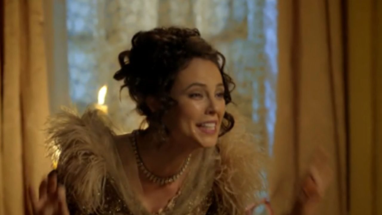 Mulher com penteado elaborado e vestido de época com detalhes de plumas sorri enquanto conversa em salão iluminado por velas em cena de novela histórica