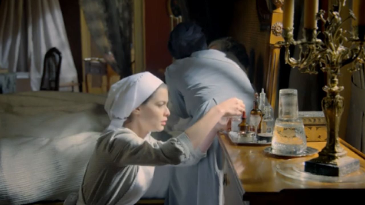 Mulher com traje de época prepara frascos sobre cômoda ao lado de cama em cena de novela histórica