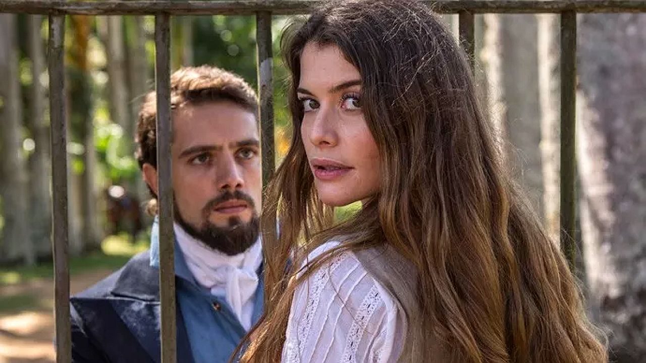 Mulher de vestido claro olha para trás diante de grade enquanto homem observa ao fundo em cena de novela de época