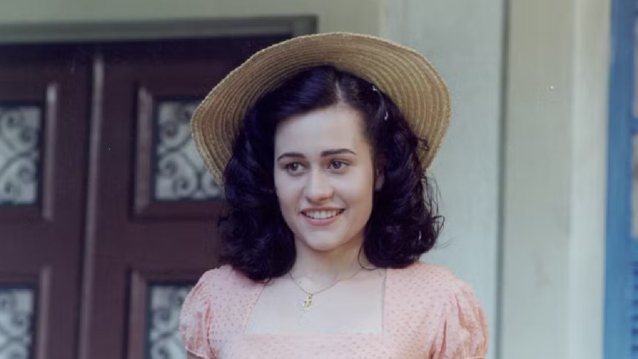 Mulher jovem com cabelos escuros cacheados, usando chapéu de palha e vestido rosa claro, sorri suavemente enquanto está de pé diante de uma porta de madeira com detalhes decorativos, em uma cena iluminada por luz natural.