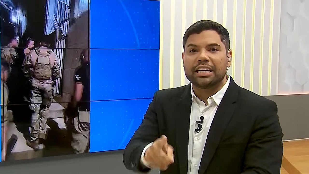Alex Silvestre aponta para câmera em estúdio enquanto comenta operação policial exibida em telão ao fundo