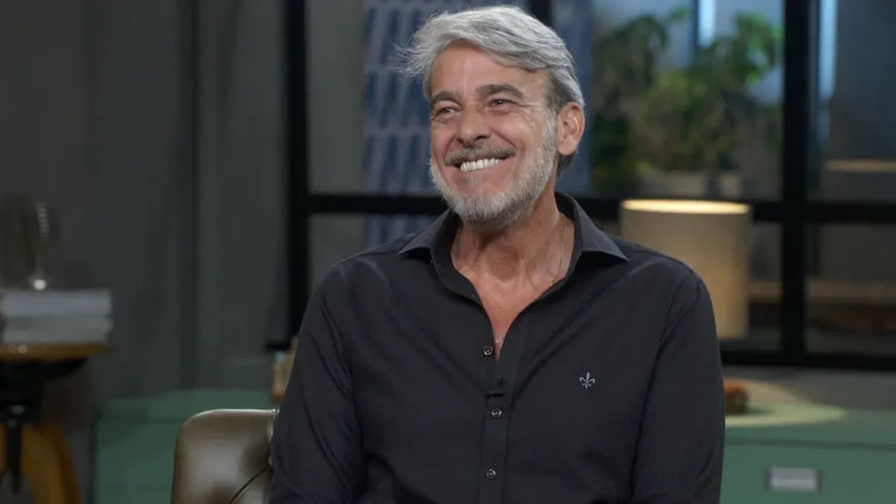 Alexandre Borges sorri durante entrevista em estúdio de televisão usando camisa preta de mangas longas