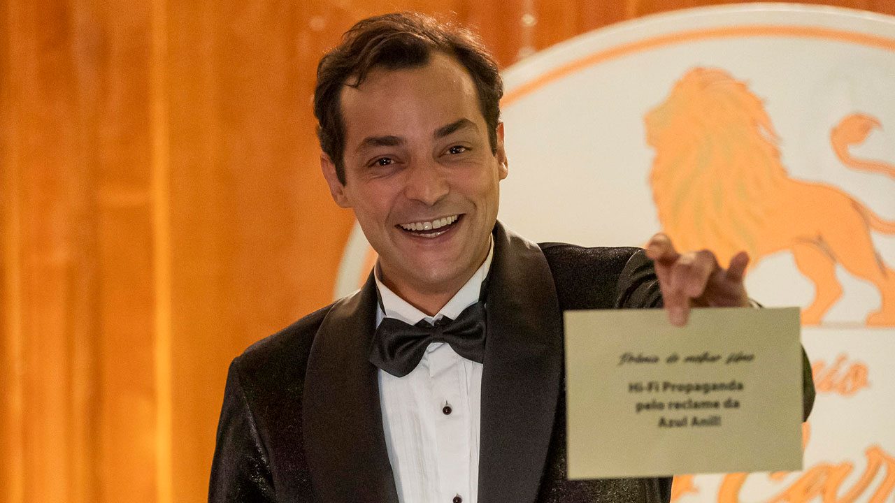 Eduardo Sterblitch caracterizado como Alfredo exibindo envelope de premiação na novela Garota do Momento