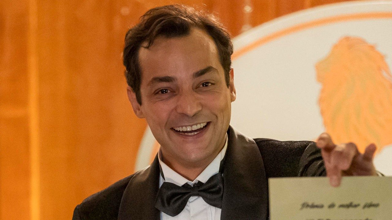 Eduardo Sterblitch como Alfredo sorri de terno e gravata borboleta segurando envelope em cena de Garota do Momento