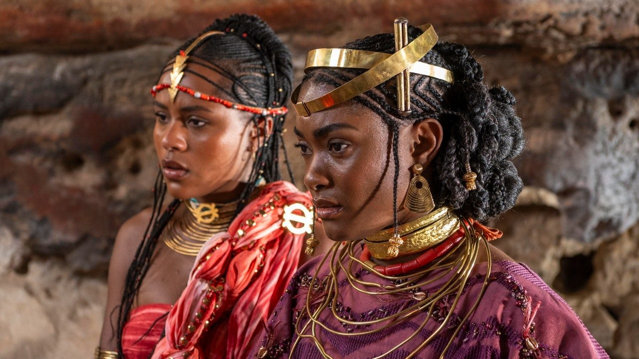 Duda Santos como Alika e Erika Januza como Niara em A Nobreza do Amor, usam penteados afro elaborados, adornos e colares dourados, roupas em tons de rosa clara