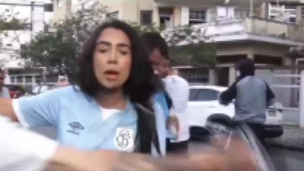 Mulher usando camisa esportiva azul clara aparece em primeiro plano em rua residencial, com expressão de fala ou protesto. Pessoas ao fundo, algumas em bicicletas, observam a cena, criando clima de movimento e tensão.