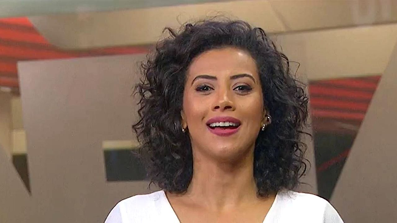 Apresentadora sorri durante programa de TV em estúdio com cenário em tons de vermelho e bege