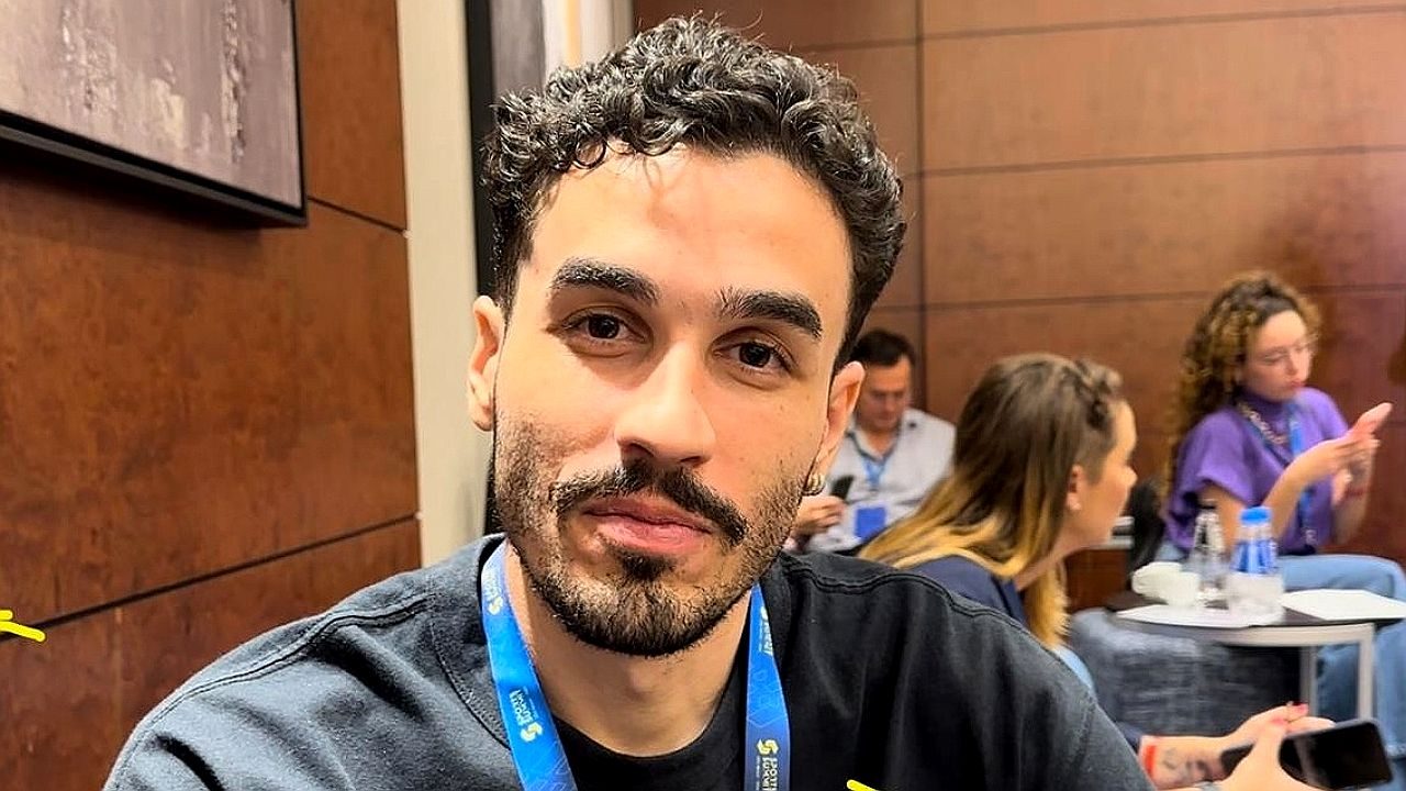 Homem com crachá azul posa durante evento em sala de conferências com pessoas ao fundo