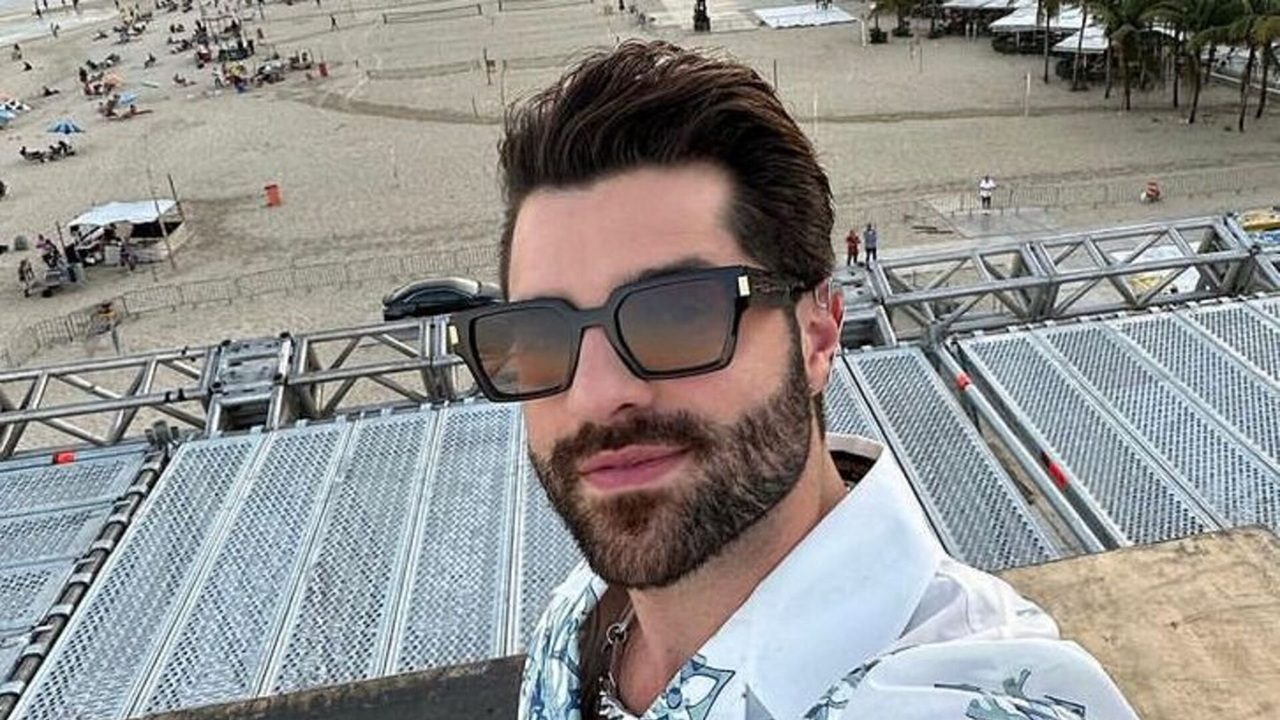 Homem jovem de barba e óculos escuros tira selfie sobre uma estrutura metálica elevada, com vista para uma praia ao fundo onde pessoas aproveitam a areia e o mar em um fim de tarde claro.