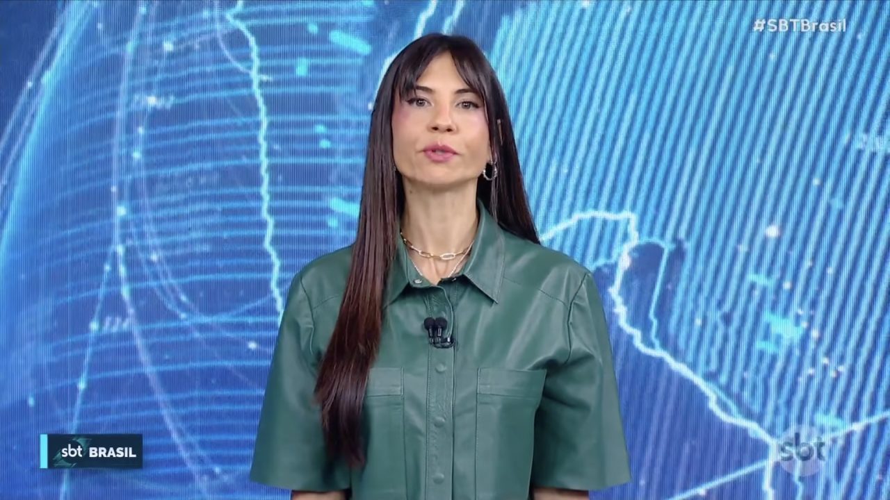 Amanda Klein, de blusa verde com microfone de lapela, diante de painel gráfico azul no estúdio do SBT Brasil