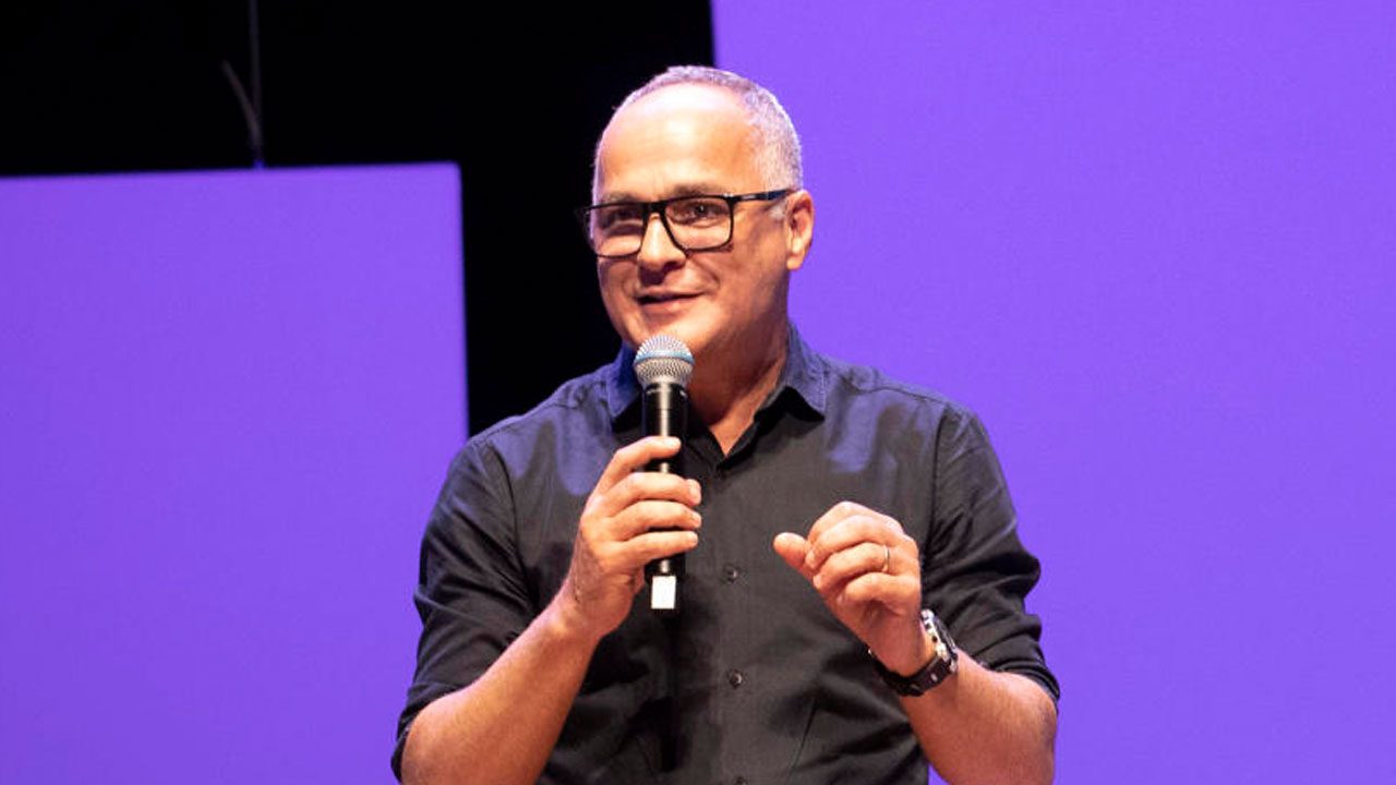 Amauri Soares fala ao microfone durante evento com fundo roxo iluminado