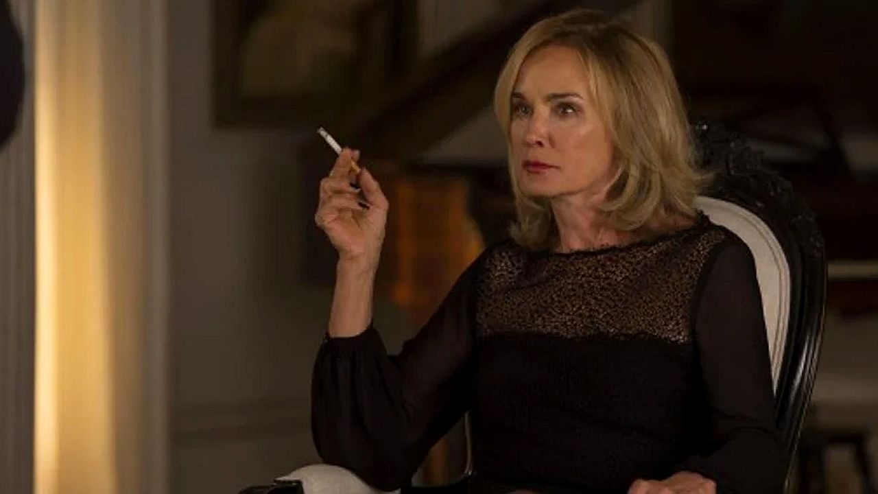 Mulher loira segura cigarro e encara alguém com expressão séria em cena de novela ambientada em sala de estar