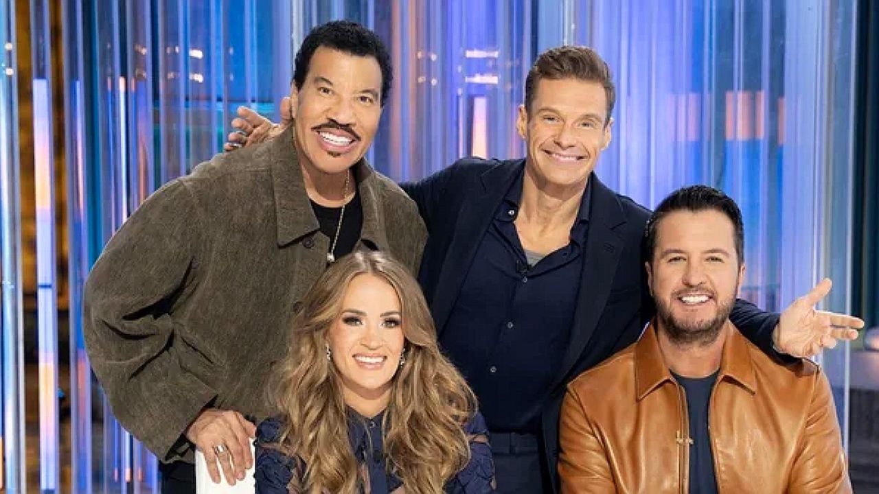 Dois homens e duas mulheres sorrindo juntos em estúdio de programa de TV com cenário iluminado ao fundo