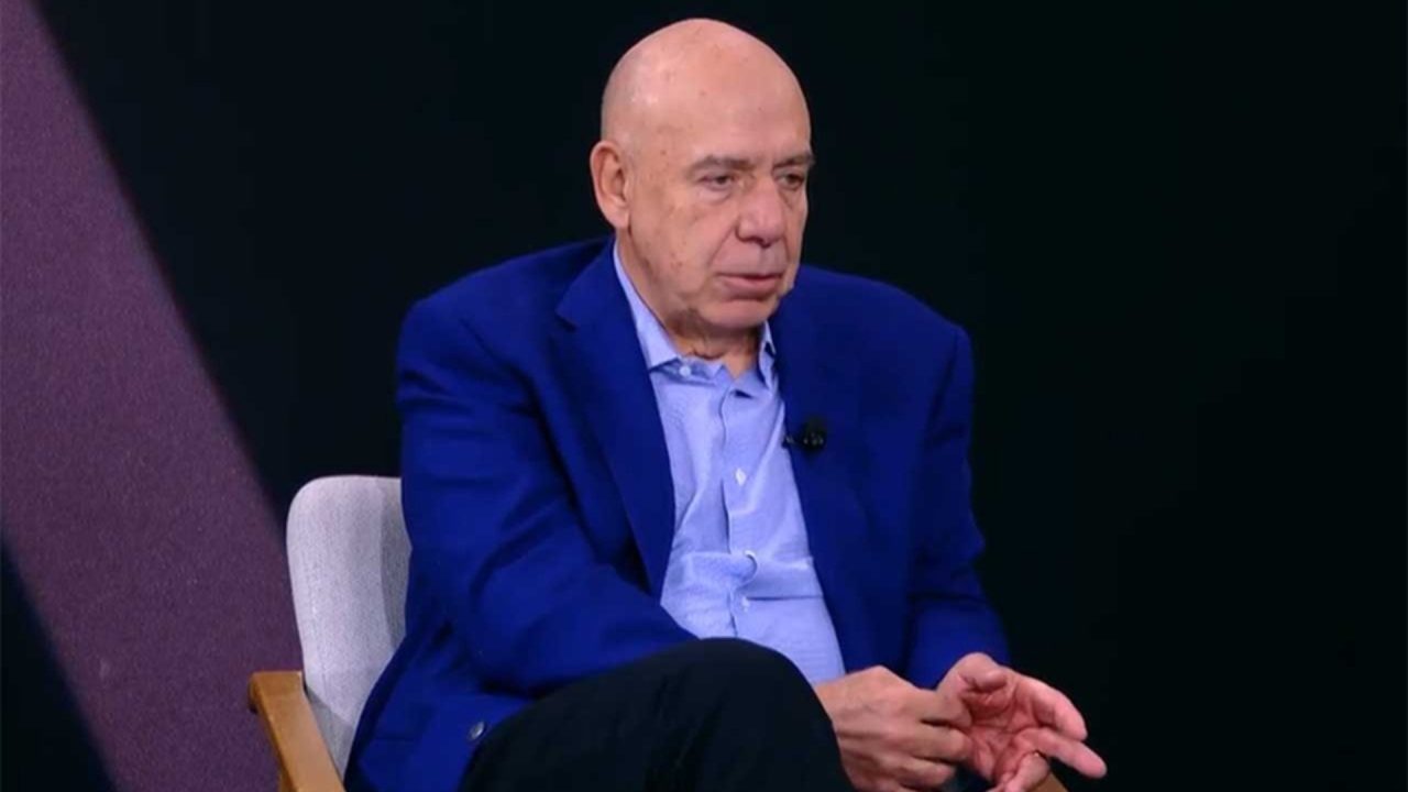 Amilcare Dallevo Jr., de terno azul, fala durante entrevista em programa da RedeTV! com fundo escuro em estúdio minimalista.