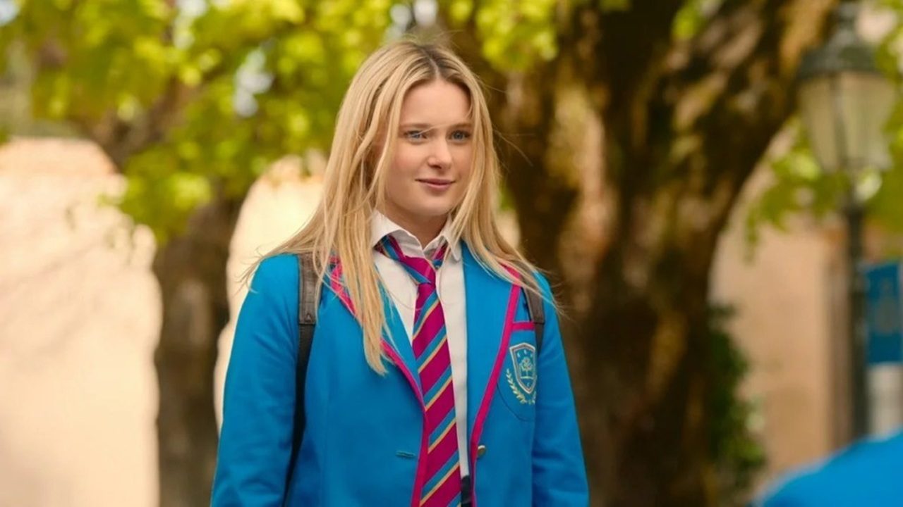 Adolescente loira veste uniforme escolar composto por blazer azul com detalhes em rosa, camisa branca e gravata listrada em tons de rosa e azul. Ela carrega mochila nas costas e está ao ar livre, diante de árvores e muro claro, com expressão atenta enquanto olha à frente.