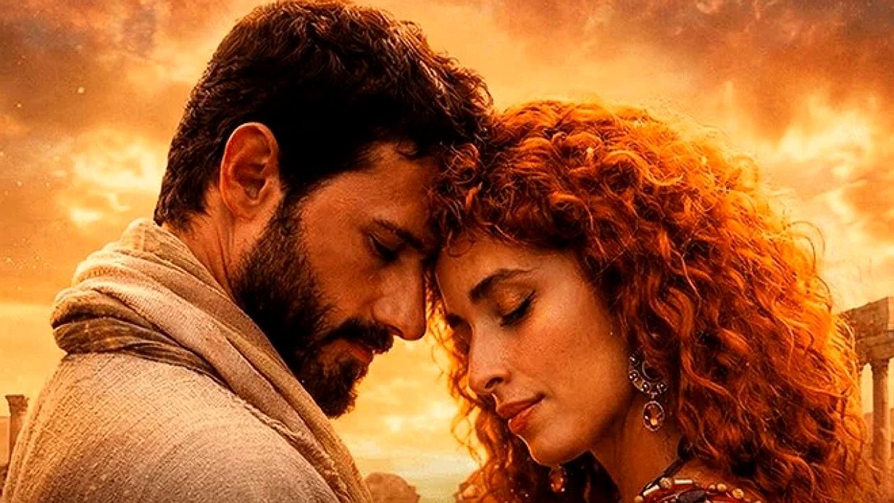 Casal com roupas de época encosta as testas em momento romântico em cenário com céu alaranjado ao fundo