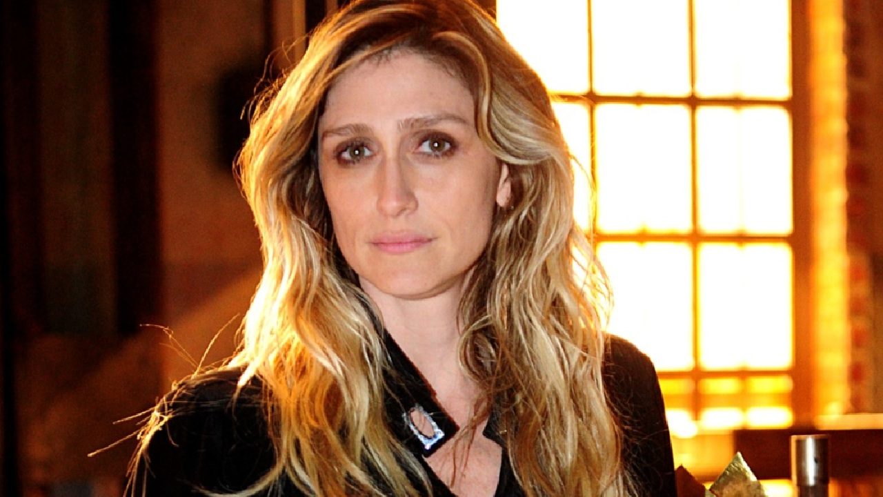 Amora Mautner com expressão séria posa em ambiente interno com iluminação quente e janela ao fundo