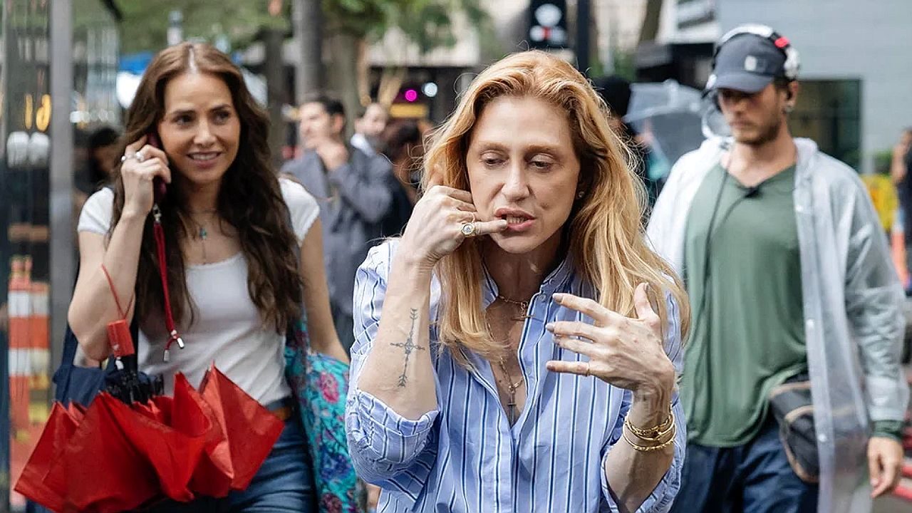 Mulher faz gesto de telefone com a mão enquanto caminha na rua acompanhada por outra mulher segurando guarda-chuva