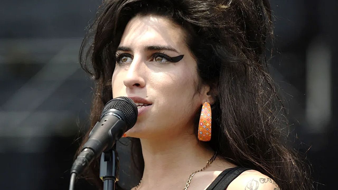 Cantora de cabelos longos e escuros se apresenta ao ar livre, cantando ao microfone. Ela usa delineado preto marcante estilo retrô, brincos grandes alaranjados e colar fino. A imagem é um close no rosto da artista, que tem expressão intensa e concentrada durante a performance.