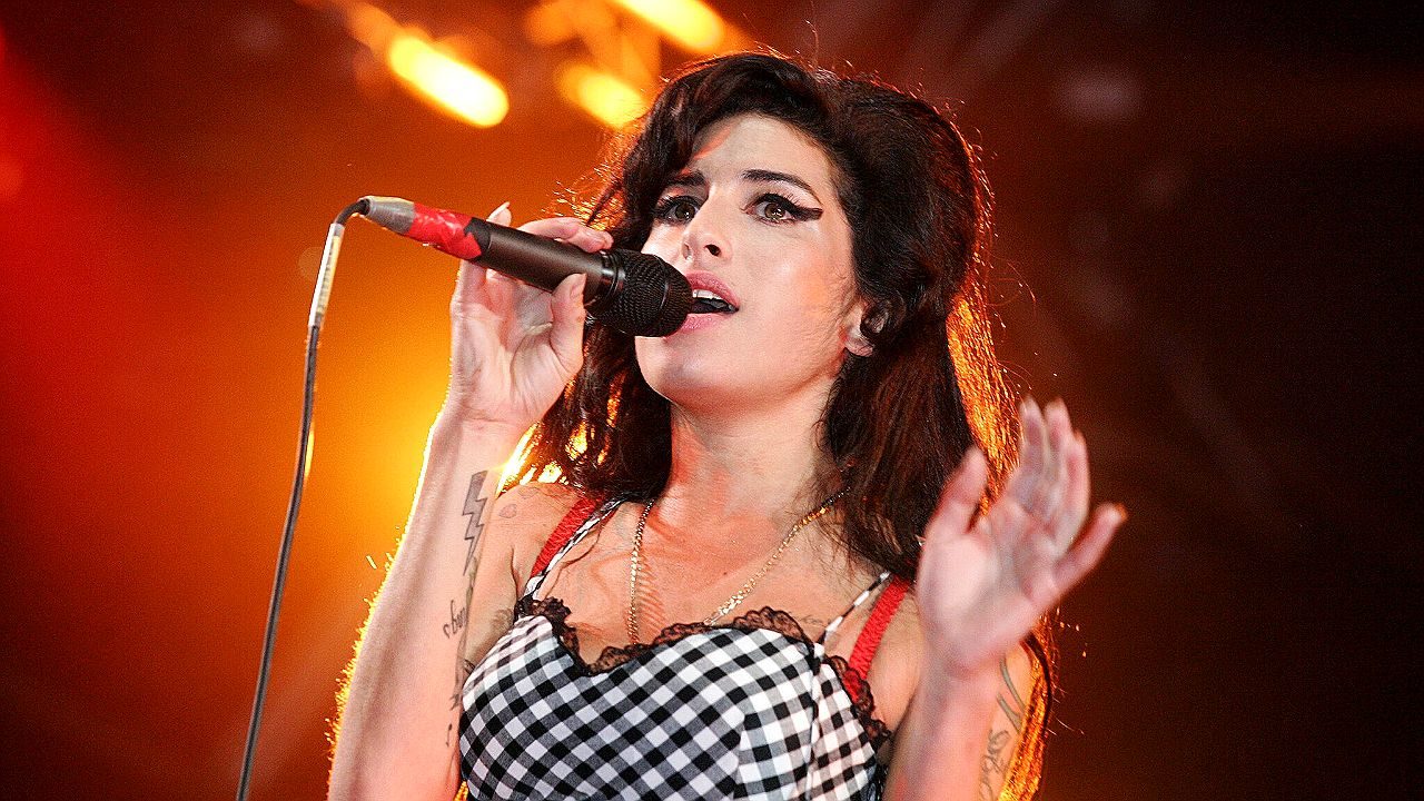 Amy Winehouse canta ao microfone em show ao vivo com iluminação quente no palco