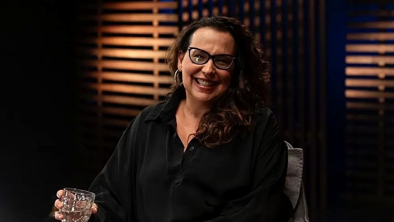 Mulher de óculos sorri durante entrevista em programa de TV com iluminação baixa no estúdio