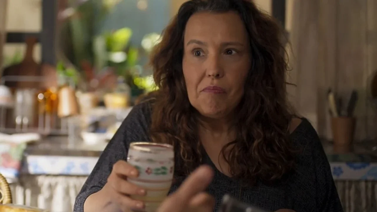 Ana Beatriz Nogueira segura copo em cena de Mania de Você, sentada à mesa em ambiente doméstico