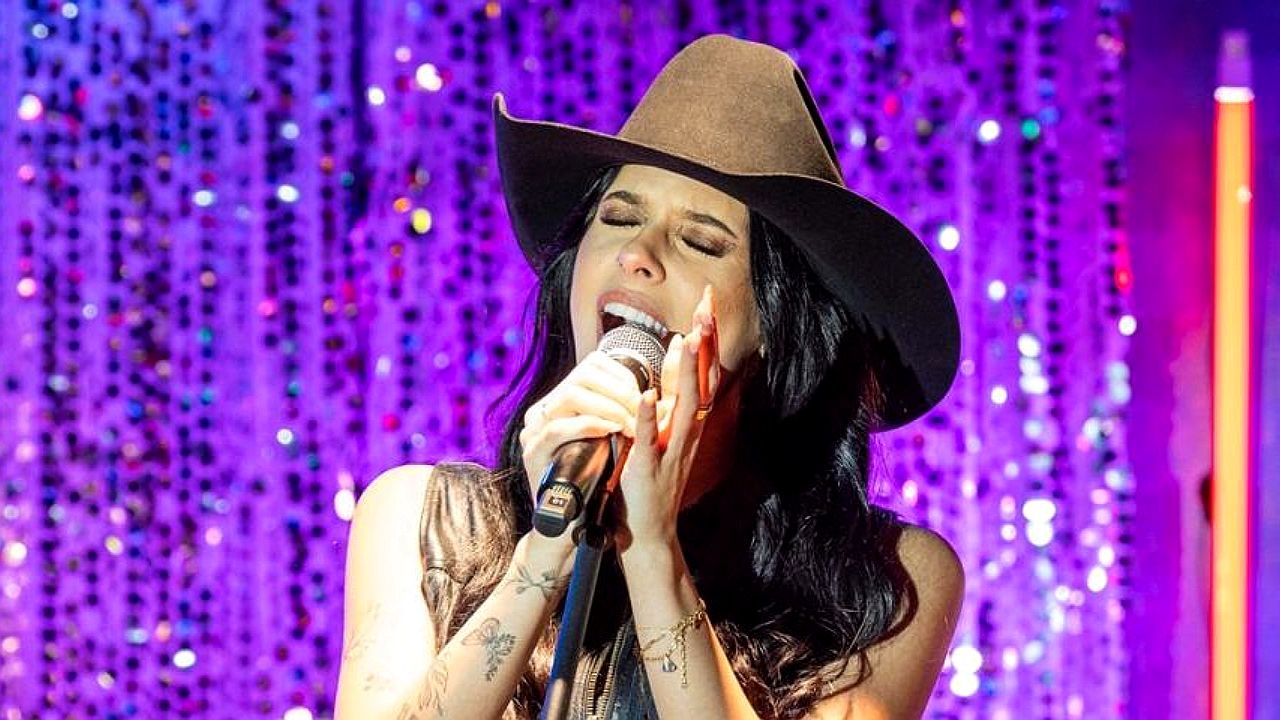Ana Castela canta ao microfone usando chapéu em apresentação com iluminação colorida ao fundo
