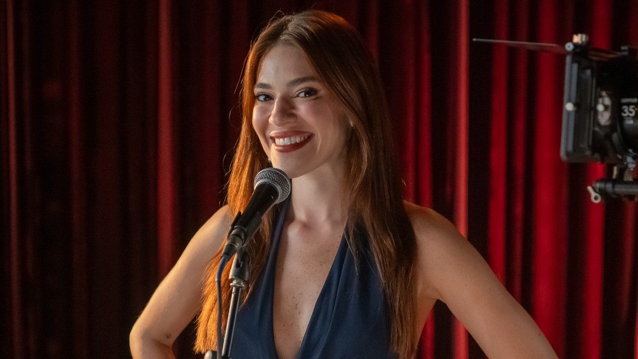 Ana Clara sorri diante de microfone em palco com cortinas vermelhas ao fundo durante gravação com câmera posicionada ao lado