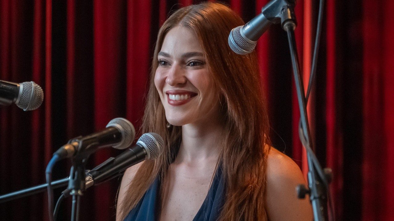 Ana Clara sorrindo diante de microfones em palco com cortinas vermelhas durante chamada do Estrelas da Casa