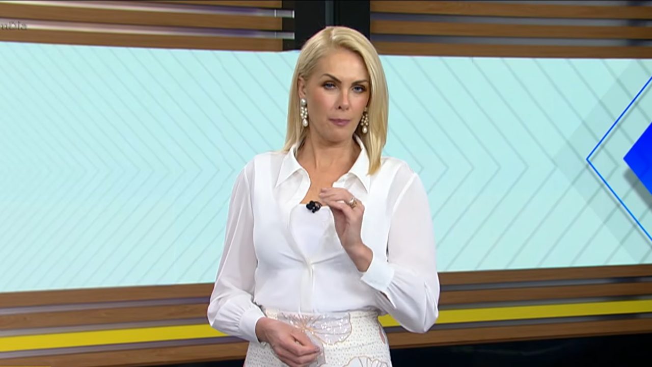 Ana Hickmann, uma apresentadora alta e loira, usa blusa branca e fala no estúdio do Hoje em Dia, fazendo gesto com a mão, diante de um painel com padrões geométricos em tons claros.