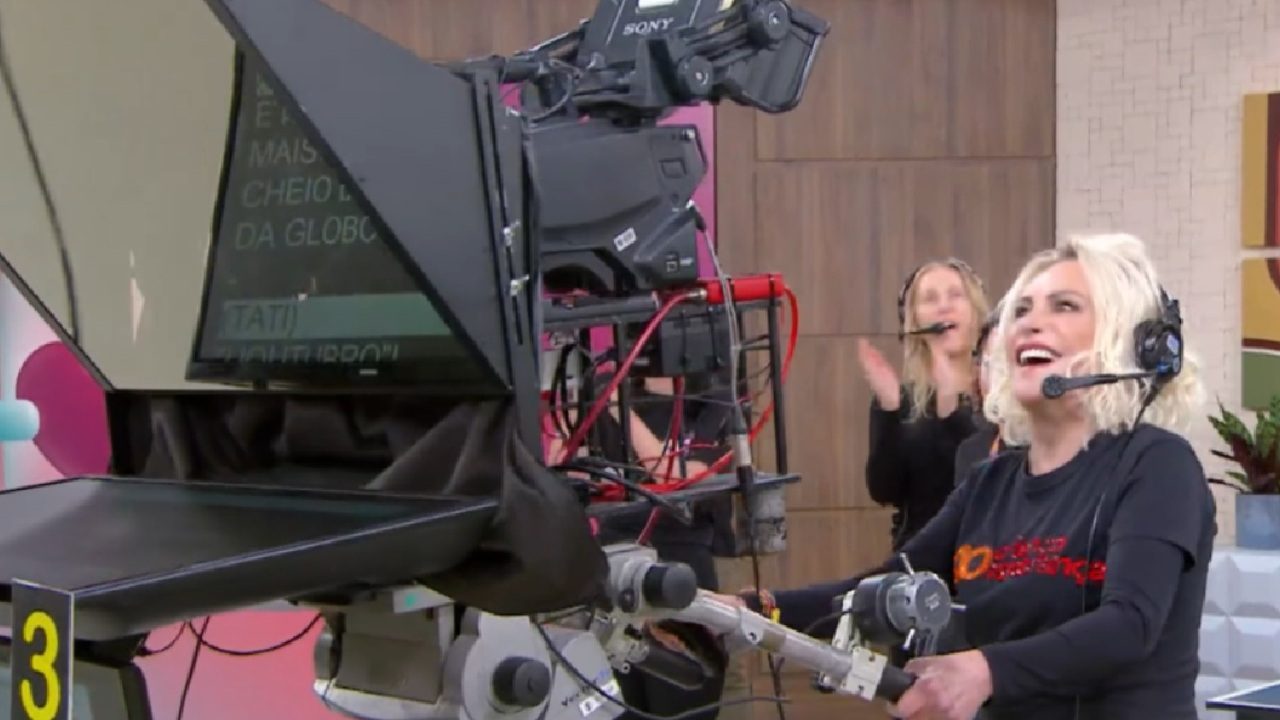 Ana Maria Braga sorridente e usando fones de ouvido com microfone, está operando uma câmera de estúdio em um ambiente de gravação com teleprompter visível e equipe ao fundo