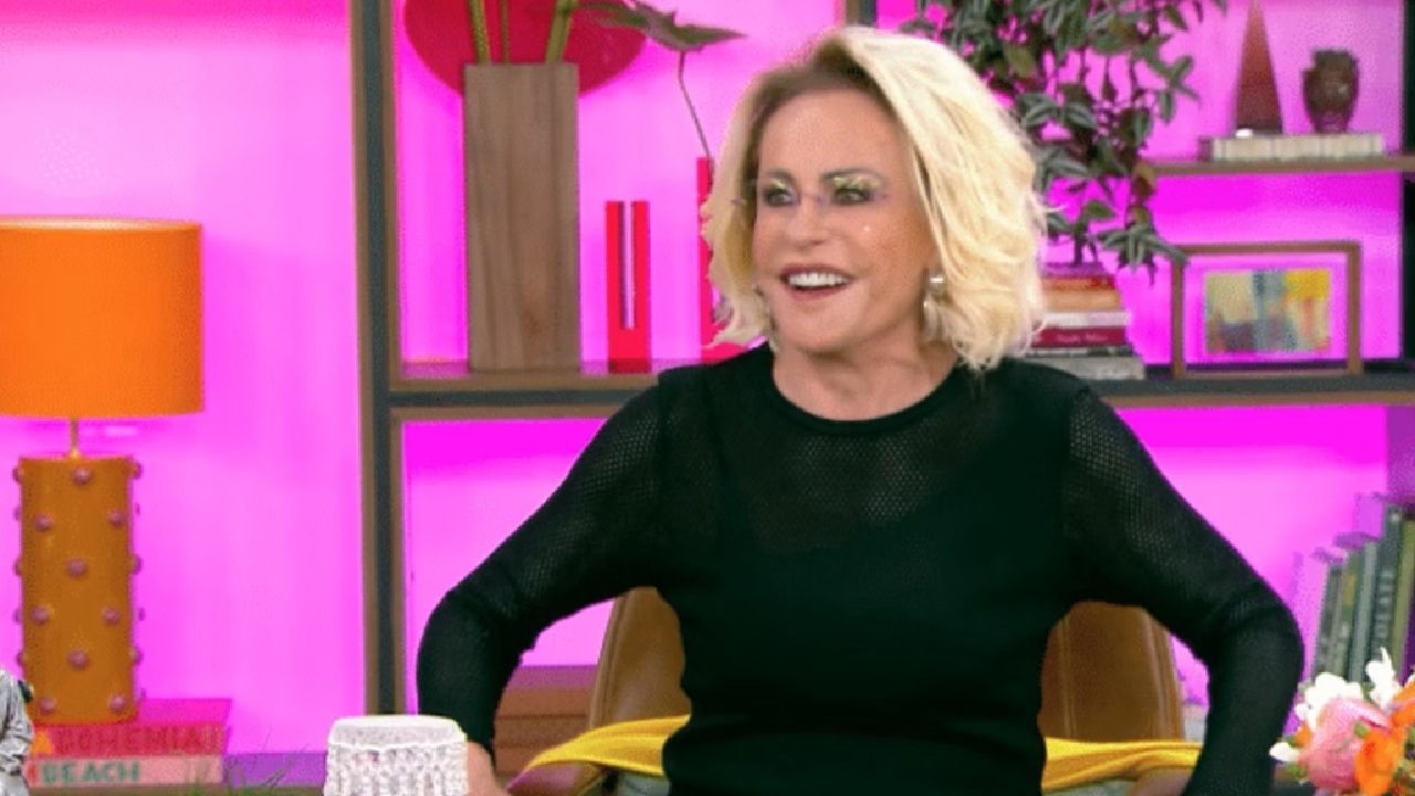 Mulher de cabelos loiros curtos sorri enquanto participa de um programa de TV em estúdio colorido. Ela veste blusa preta de tecido vazado e está sentada em uma poltrona, com as mãos apoiadas nos braços. Ao fundo, há prateleiras com livros, plantas e objetos decorativos sobre uma parede iluminada em rosa vibrante.