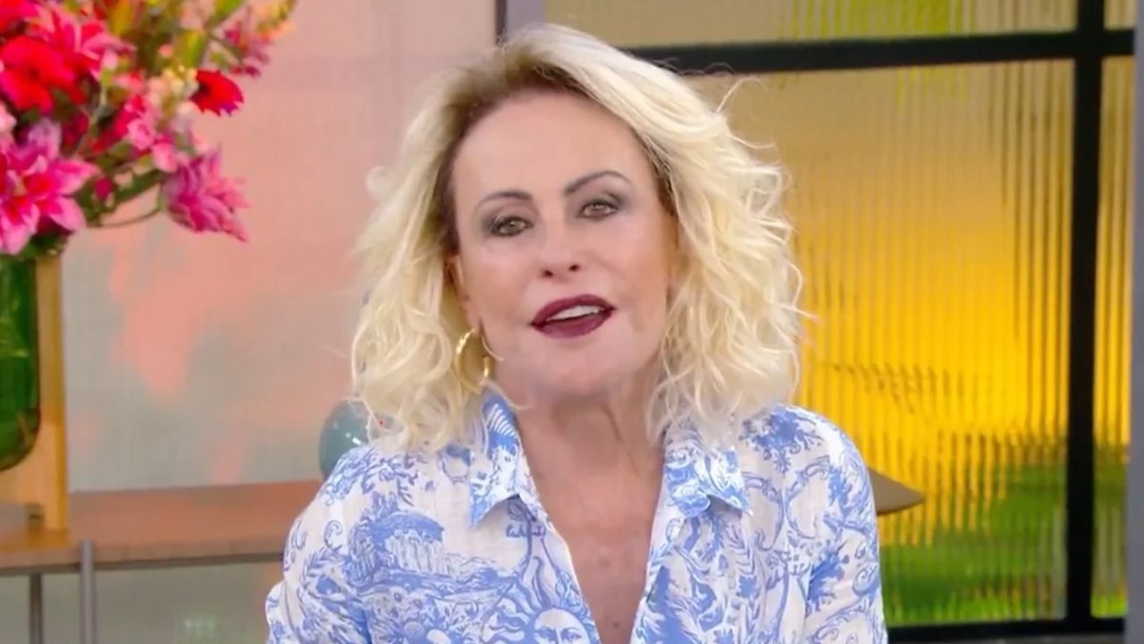 Apresentadora de cabelos loiros cacheados, usando camisa estampada em azul e branco, aparece sorrindo em um estúdio iluminado. Ao fundo, há flores vibrantes em tons de rosa e elementos decorativos com luz amarelada, criando um clima acolhedor de programa matinal.