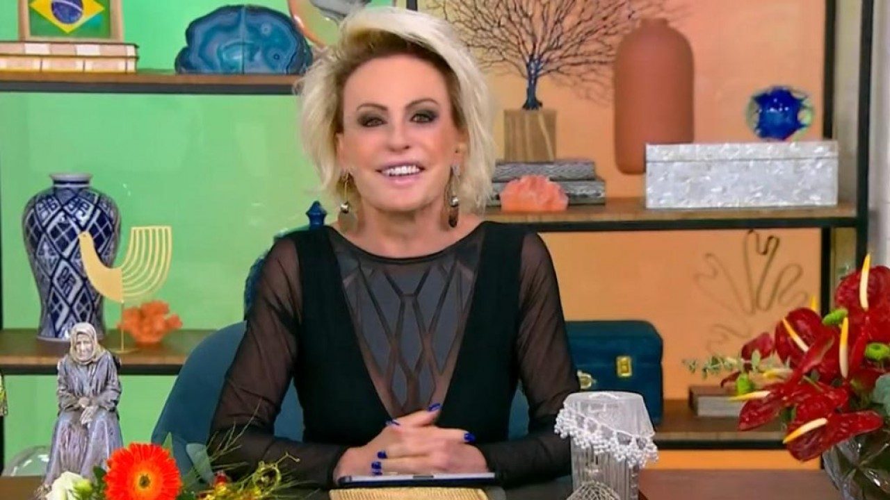 Apresentadora sentada à mesa sorri para a câmera em um estúdio colorido decorado com vasos, flores, esculturas e objetos artesanais. Ela usa blusa preta com mangas transparentes e tem maquiagem marcada, enquanto apoia as mãos sobre a mesa ornamentada com arranjos florais