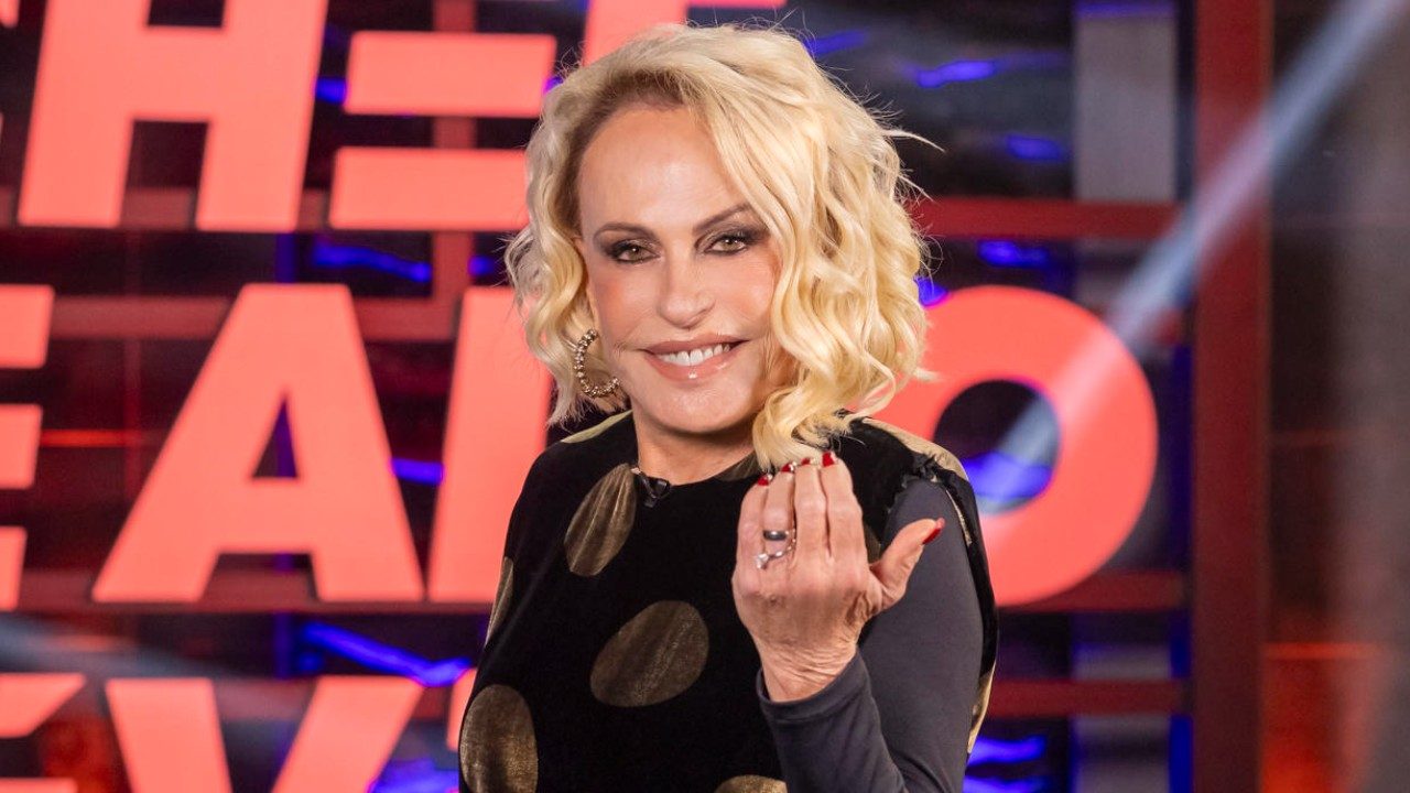 Ana Maria Braga sorri e faz gesto com a mão em estúdio usando vestido preto com círculos dourados e fundo iluminado