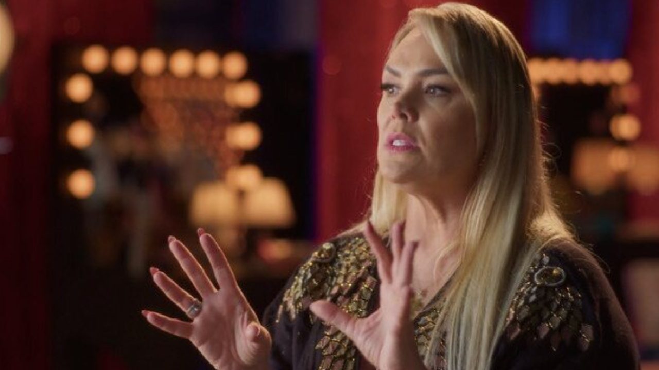 A imagem mostra uma mulher loira de cabelos longos, gesticulando com as mãos enquanto fala. Ela veste uma blusa escura com detalhes brilhantes nos ombros e está em um ambiente com iluminação suave e espelhos rodeados por luzes, sugerindo um camarim ou estúdio de gravação