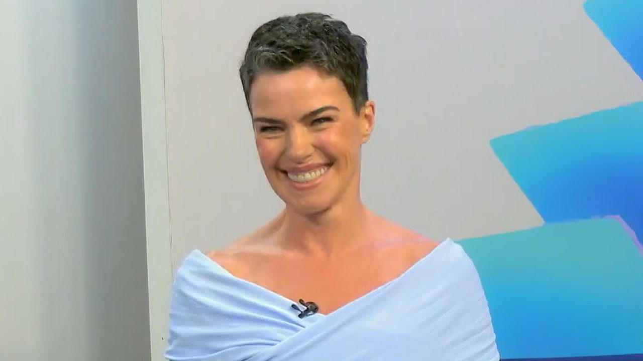 Ana Paula Arósio sorrindo com blusa azul claro e microfone preso na roupa durante entrevista