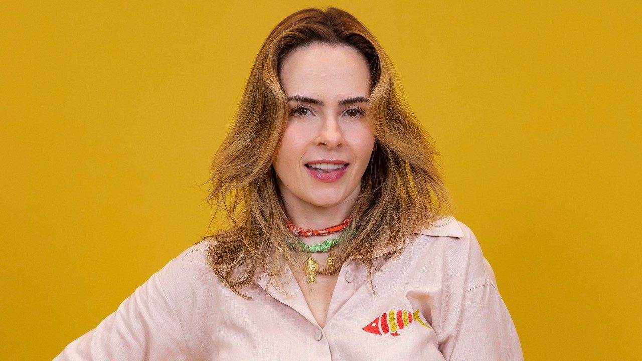 Ana Paula Renault de camisa bege com estampa de peixe colorido e colares artesanais posa sorridente em frente a um fundo amarelo vibrante e liso