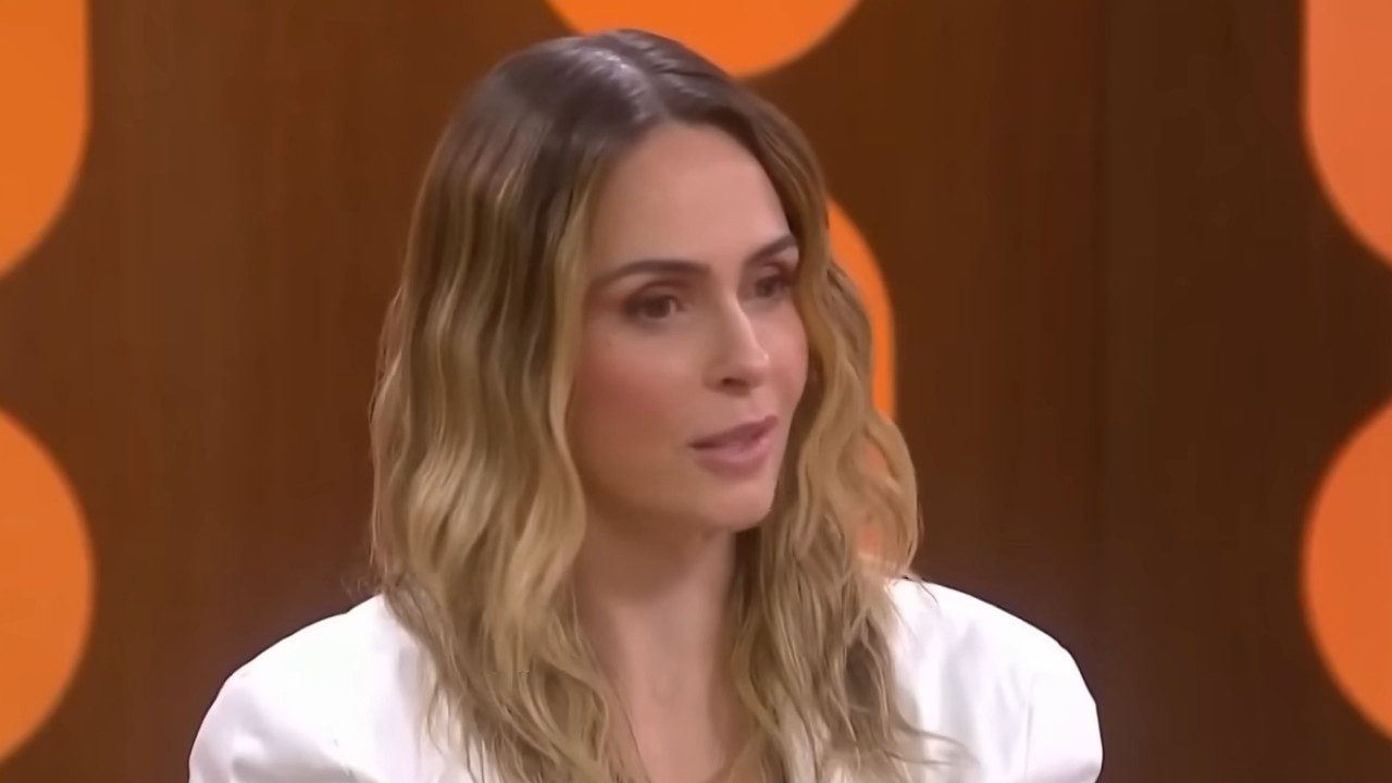 Ana Paula Renault durante participação no Saia Justa com blazer branco e cenário iluminado ao fundo