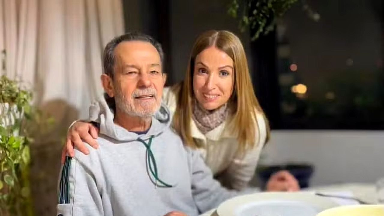 Ananda Apple posa ao lado de homem sentado à mesa durante encontro em ambiente residencial iluminado à noite