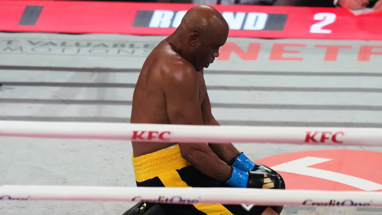 Lutador de boxe ajoelhado no ringue com luvas azuis e calção amarelo durante combate sob iluminação de arena esportiva